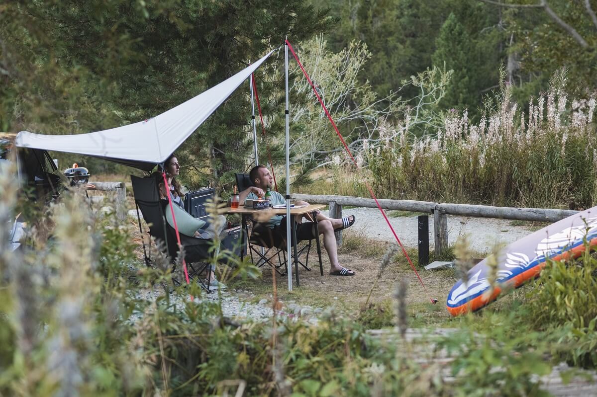 3 Freunde sitzen um einen Campingtisch unter einem Dachzelt vor dem Auto im Sommer in der natur