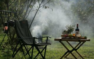 hübsch mit Wein, Früchten und Kräutertopf gedeckter Campingtisch mit Campingstuhl im Grünen vor rauchendem Feuer