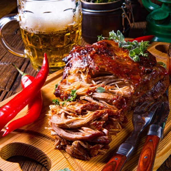 Pulled Pork auf heller Holzplatte mit roten Chillies dekoriert und Besteck auf dunklem Holztisch mit Bierglas mit Henkel im Hintergrund