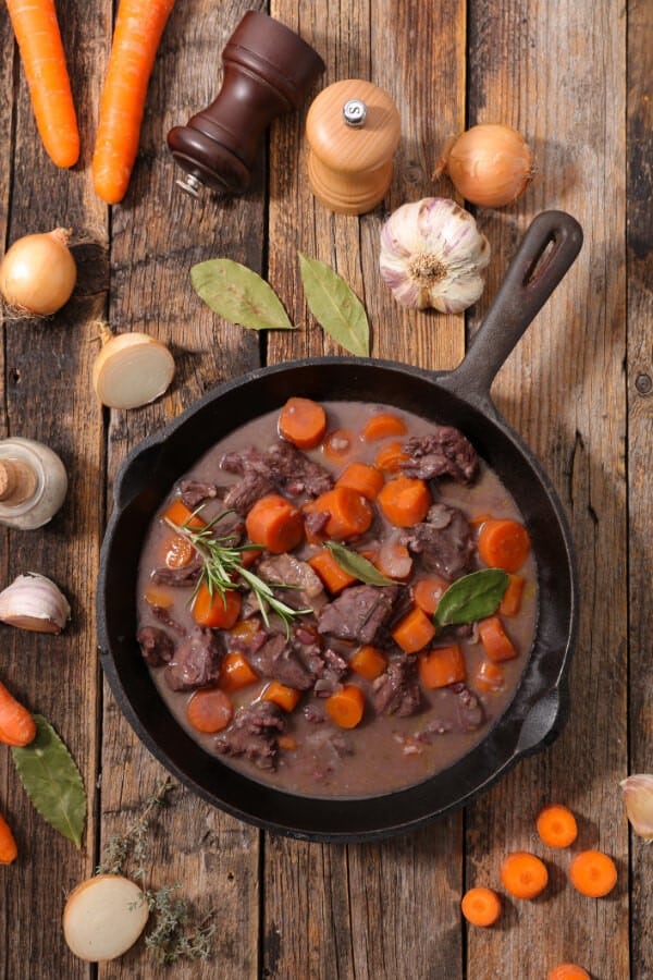 Rindergulasch mit Möhrenstückchen in einer schwarzen Gussplfanne auf einem Holztisch, der mit Knoblauch, Zwiebelhälften, Salz- und Pfefferstreuer und Lobeerblättern dekoriert ist