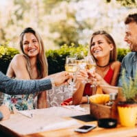 4 Freunde sitzen draußen an einem Holztisch in einem Gartenrestaurant im Sommer und prosten sich mit Weißweingläsern zu.