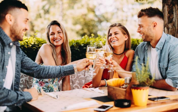 4 Freunde sitzen draußen an einem Holztisch in einem Gartenrestaurant im Sommer und prosten sich mit Weißweingläsern zu.
