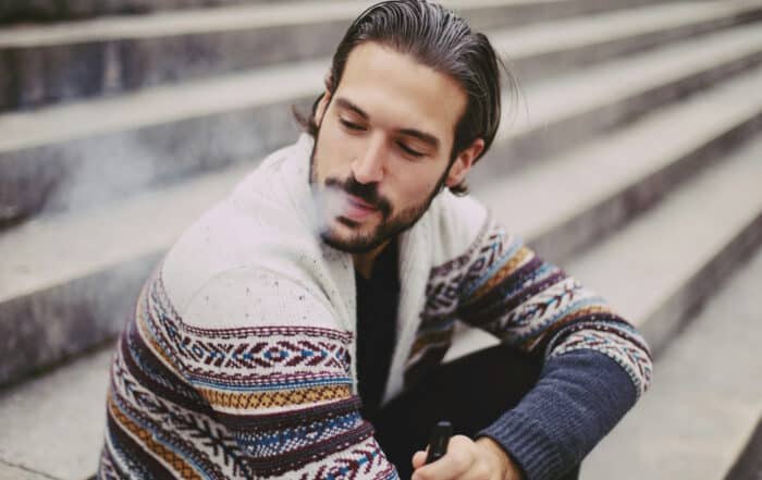 Junger Mann sitzt auf einer Treppe im Freien und raucht eine E-Zigarette