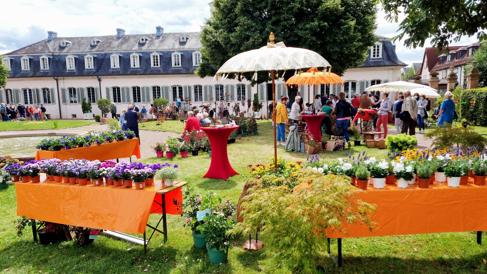 Genuss- und Gartenfest auf Schloss Braunshardt Genuss- und Gartenfest auf Schloss Braunshardt - Menschen schlendern durch den Schlosspark mit Ausstellern und Blumen, Sonnenschirmen und Stehtischen mit roten Hussen.