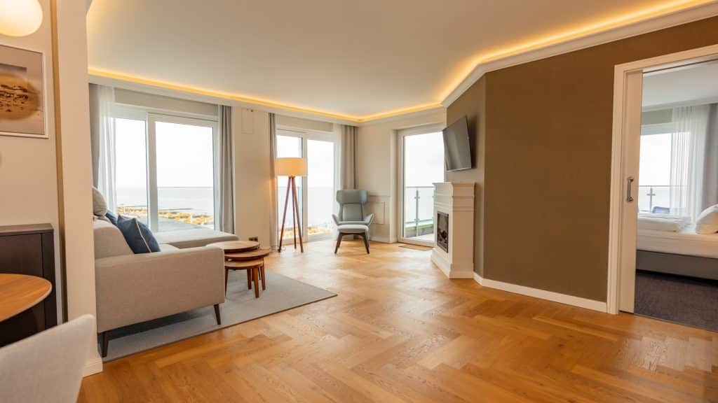 großzügige Suite mit Parkettboden Sitzecke, bodentiefen Fenstern, Kamin und Blick ins Schlafzimmer im Hotel Strandperle Duhnen