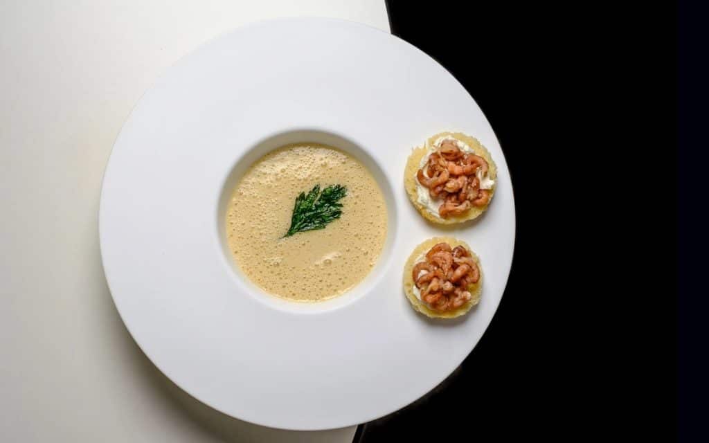 luxus-gourmet-rezept-krabbensuppe-thomas-sixt-fuer-varta Krabbensuppe mit Petersilienblatt und 2 runden Kräckern mit Krabben am Tellerrand in einem weißen Suppenteller mit breitem Rand