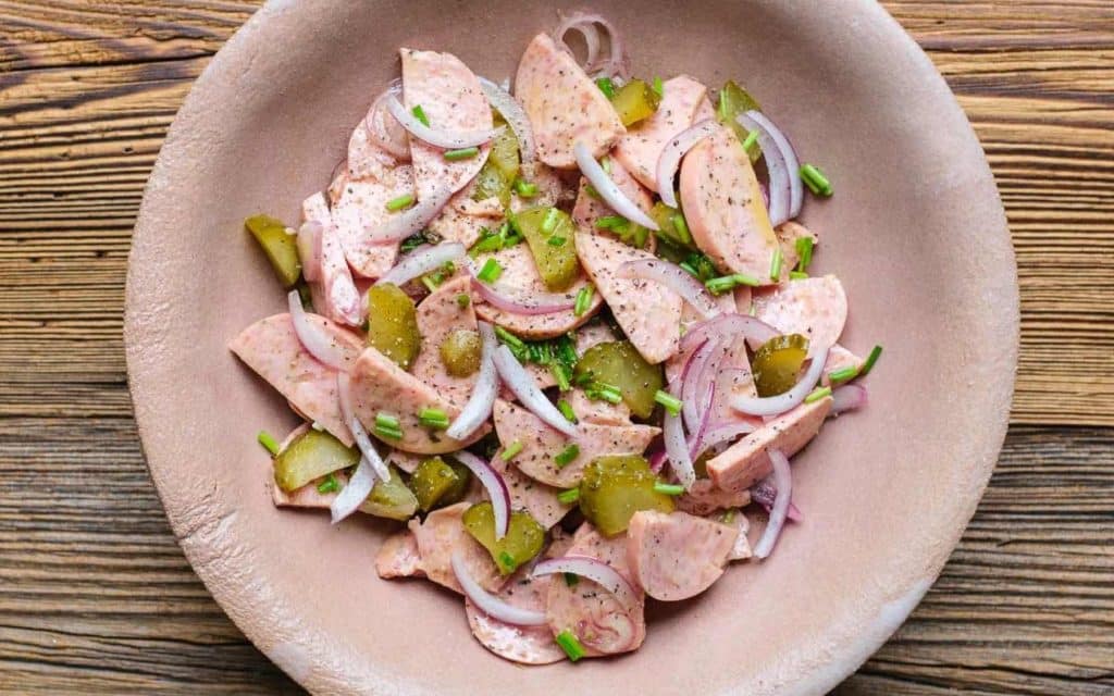 Wurstsalat mit Zwiebeln und Gurken in beigefarbenen tiefen Tonteller auf Holztisch