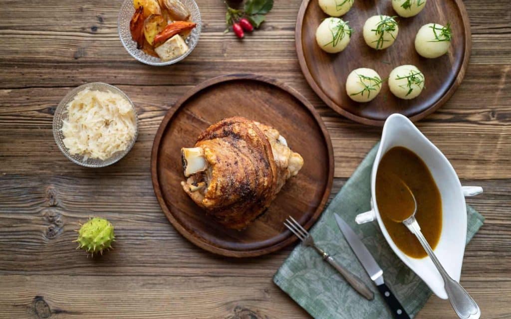 knusprige Schweinshaxe auf Holzteller, daneben Holzteller mit Semmelknödeln, Sauciere mit brauner Soße und zwei Glasschälchen mit Gemüse