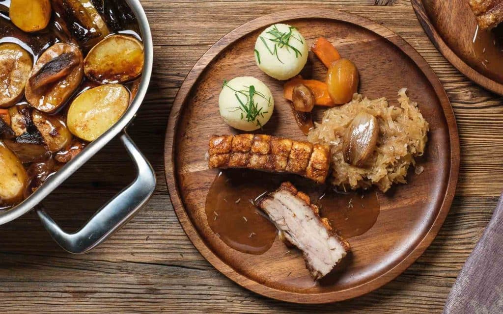 Schweinsbraten mit Semmelknödel und Kraut auf Holzteller neben Kasserrolle mit Kartoffeln