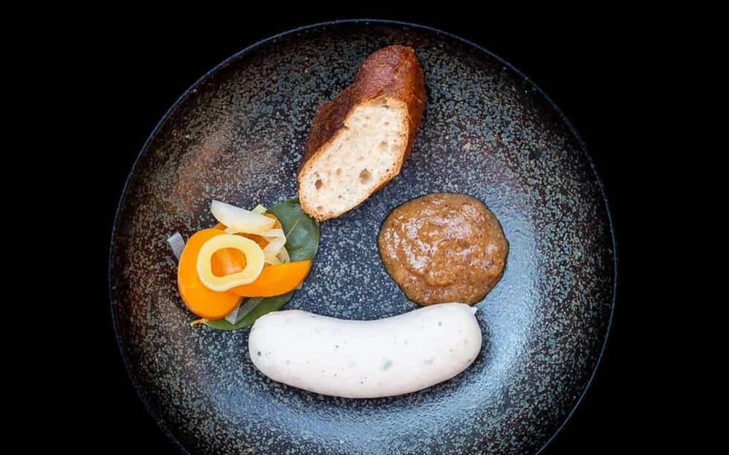 Weißwurst mit süße, Senf und Brezelstück und eingelegten Karottenscheiben auf einem schwarz-grauen Steingutteller