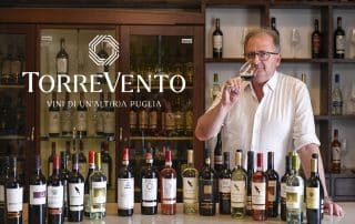 Person steht hinter einer Theke, auf der eine große Auswahl von Weinflaschen in verschiedenen Sorten und Farben aufgereiht sind. Im Hintergrund ein Regal mit weiteren Flaschen. Das Logo ‚TORREVENTO – Vini di un’altra Puglia‘ ist links neben der Person eingefügt.