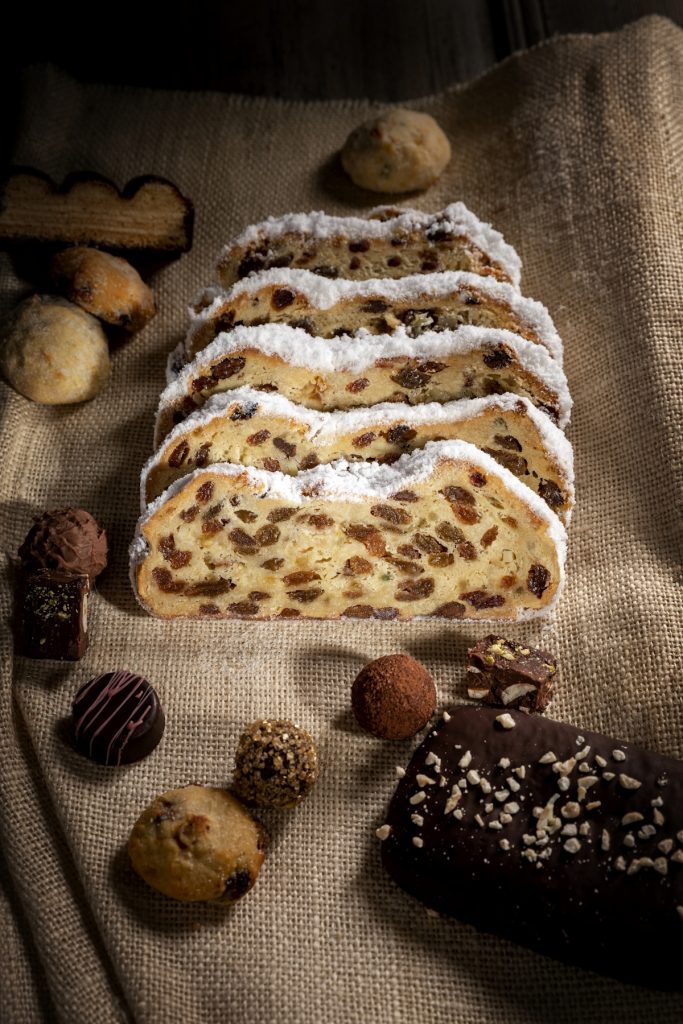 20020_Dresdner_Stollen_Frauenkirche_mood_s Mehrere Scheiben Christstollen mit Puderzucker auf einem Leinentuch, umgeben von Pralinen, Keksen und einem Schokoladengebäck.