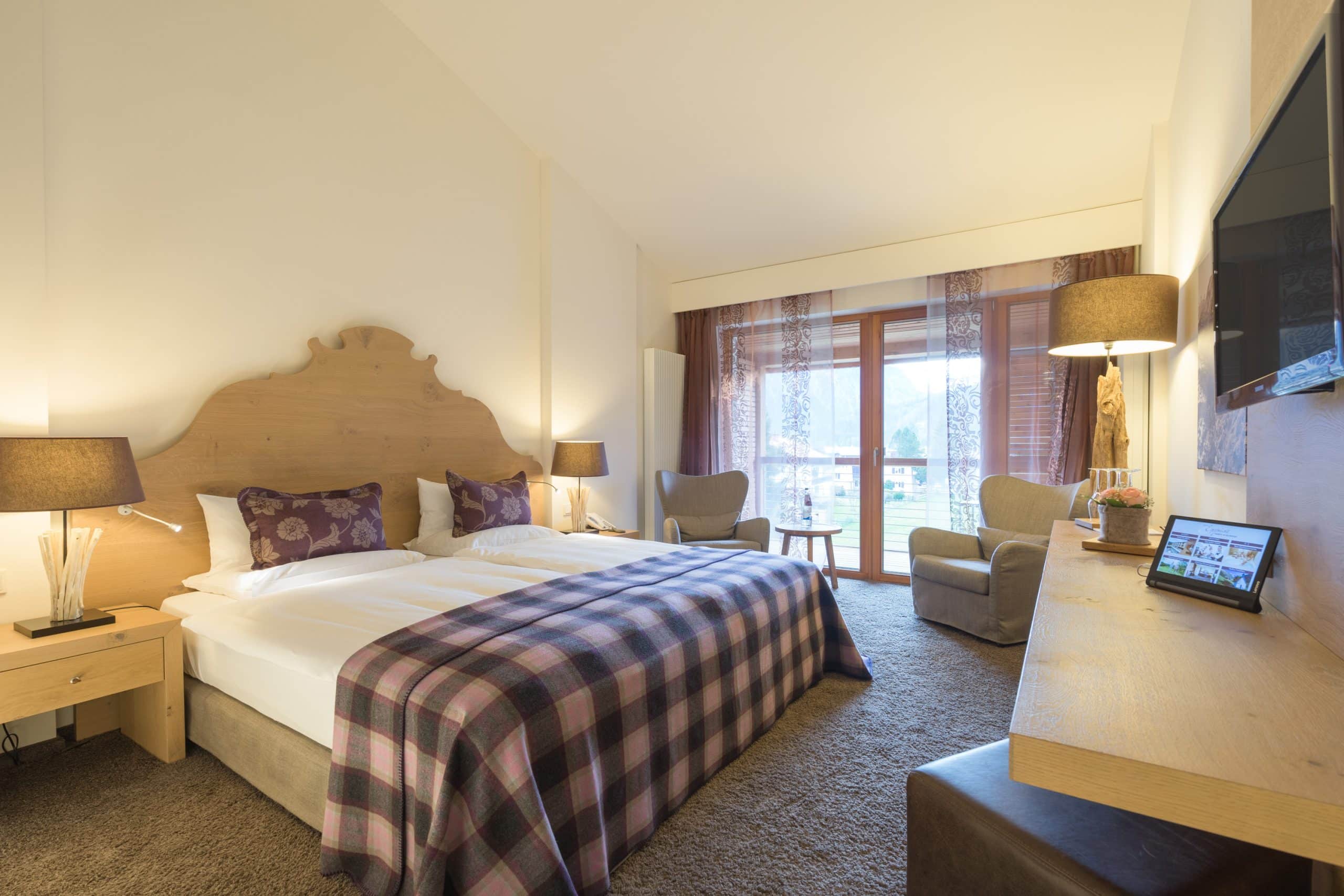 2_Exquisit Helles Hotelzimmer mit Doppelbett, zwei Sesseln, Schreibtisch und großem Fenster mit Blick auf die Berge.