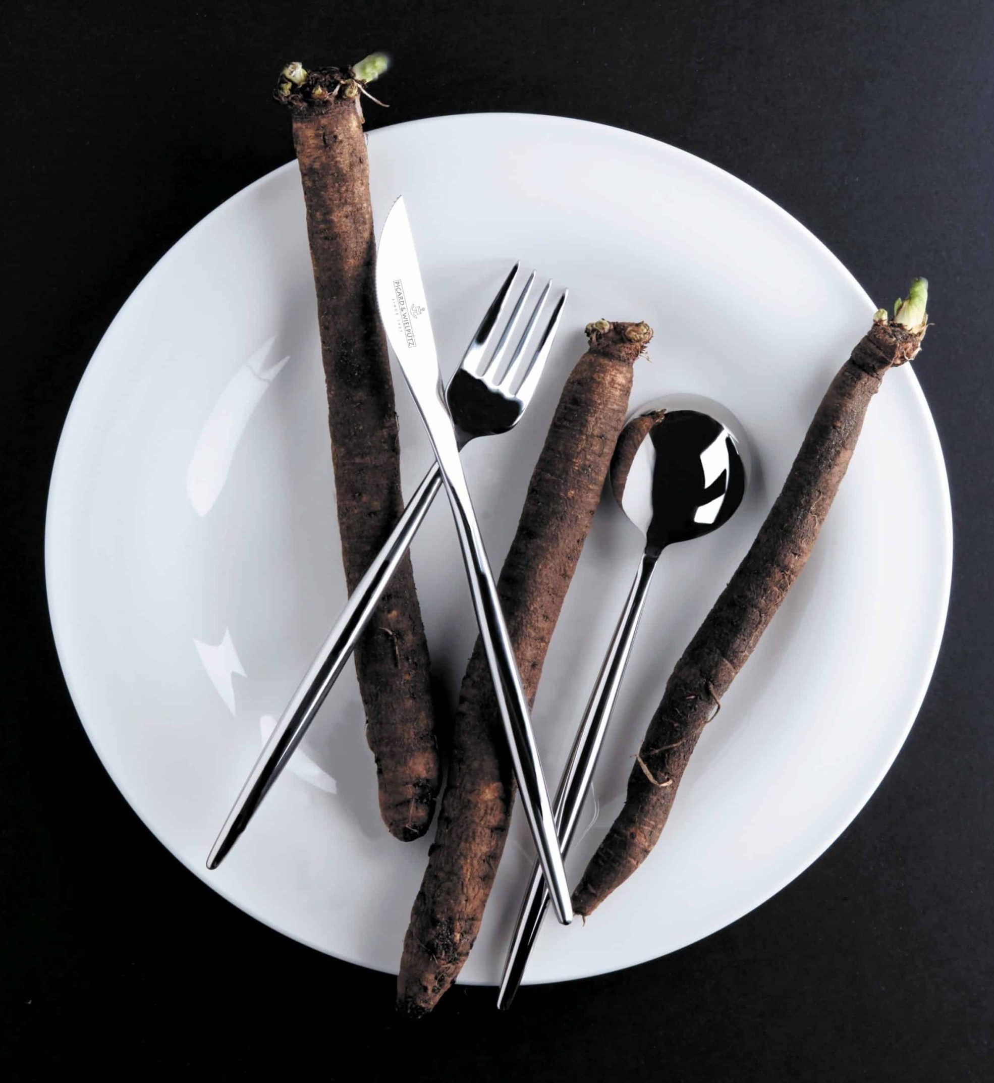 6116 Lotus-Mood-shot 1 Weiße Teller mit vier dunklen Wurzeln und drei Besteckteilen auf schwarzem Hintergrund.