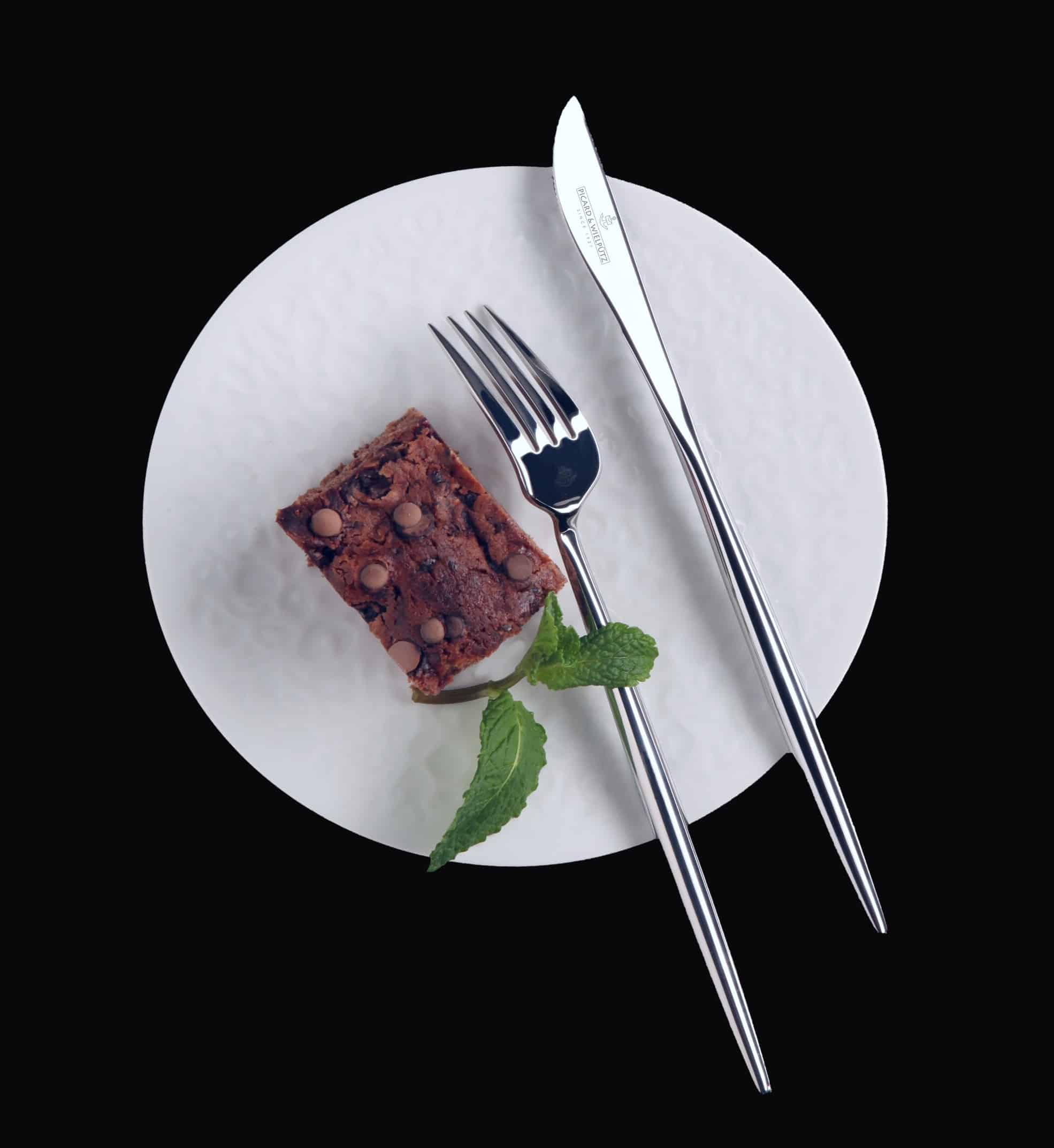 6116 Lotus-Mood-shot 2 Weißer Teller mit einem Stück Schokoladenkuchen und Minzblatt, daneben Messer und Gabel auf schwarzem Hintergrund.