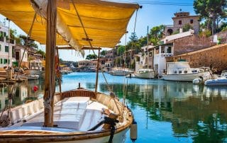 Holzsegelboot mit gelbem Sonnensegel in einem ruhigen mediterranen Hafen mit türkisfarbenem Wasser und Häusern im Hintergrund.