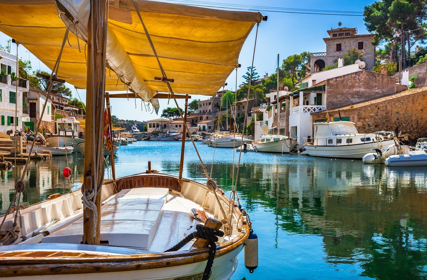 Holzsegelboot mit gelbem Sonnensegel in einem ruhigen mediterranen Hafen mit türkisfarbenem Wasser und Häusern im Hintergrund.