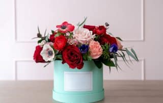 Runder, türkisfarbener Blumenbehälter auf einem Tisch, gefüllt mit einem gemischten Blumenarrangement aus roten, rosa und weißen Blumen.