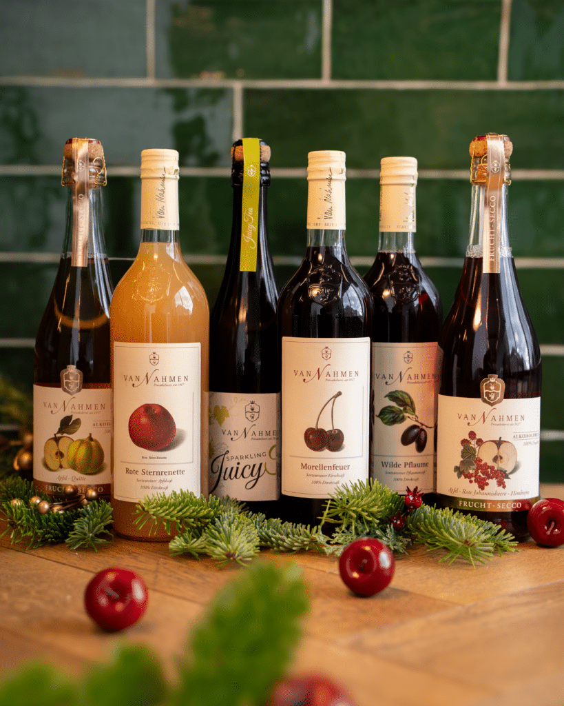 Sechs Flaschen Obstwein und Likör in verschiedenen Sorten, darunter Apfel, Zwetschge und Beeren, mit weihnachtlicher Deko.