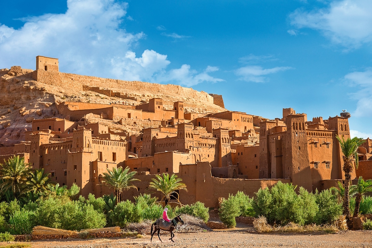 historische Wüstenstand aus Sandstein an einem Berghang im Vordergrund Palemn und grüne Sträucher und ein Reiter auf einem Esel