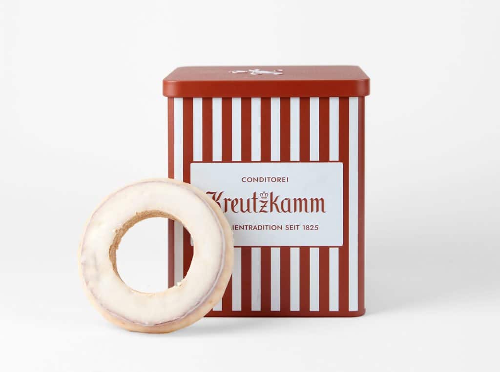 Ein rot-weiß gestreifter Metallbehälter mit der Aufschrift ‚Conditorei Kreutzkamm – Feine Tradition seit 1825‘. Daran angelehnt ein runder Gebäckring mit weißer Glasur.