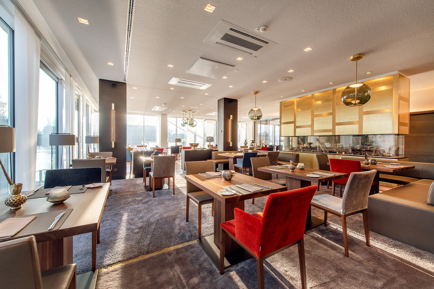 H16246mp_Gastronomie_media_image_1 Helles, modernes Restaurant mit Holztischen, roten und grauen Polsterstühlen sowie großen Fenstern.