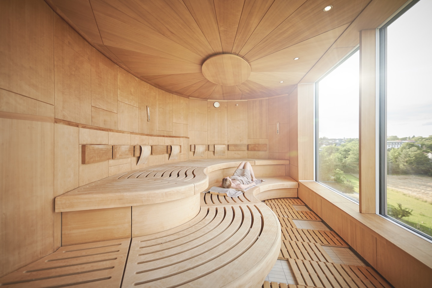 H16246mp_Wellness_media_image_3 Große Sauna aus hellem Holz mit breiten Sitzflächen und Panoramafenster mit Blick ins Grüne. Eine Person liegt auf der unteren Sitzfläche.