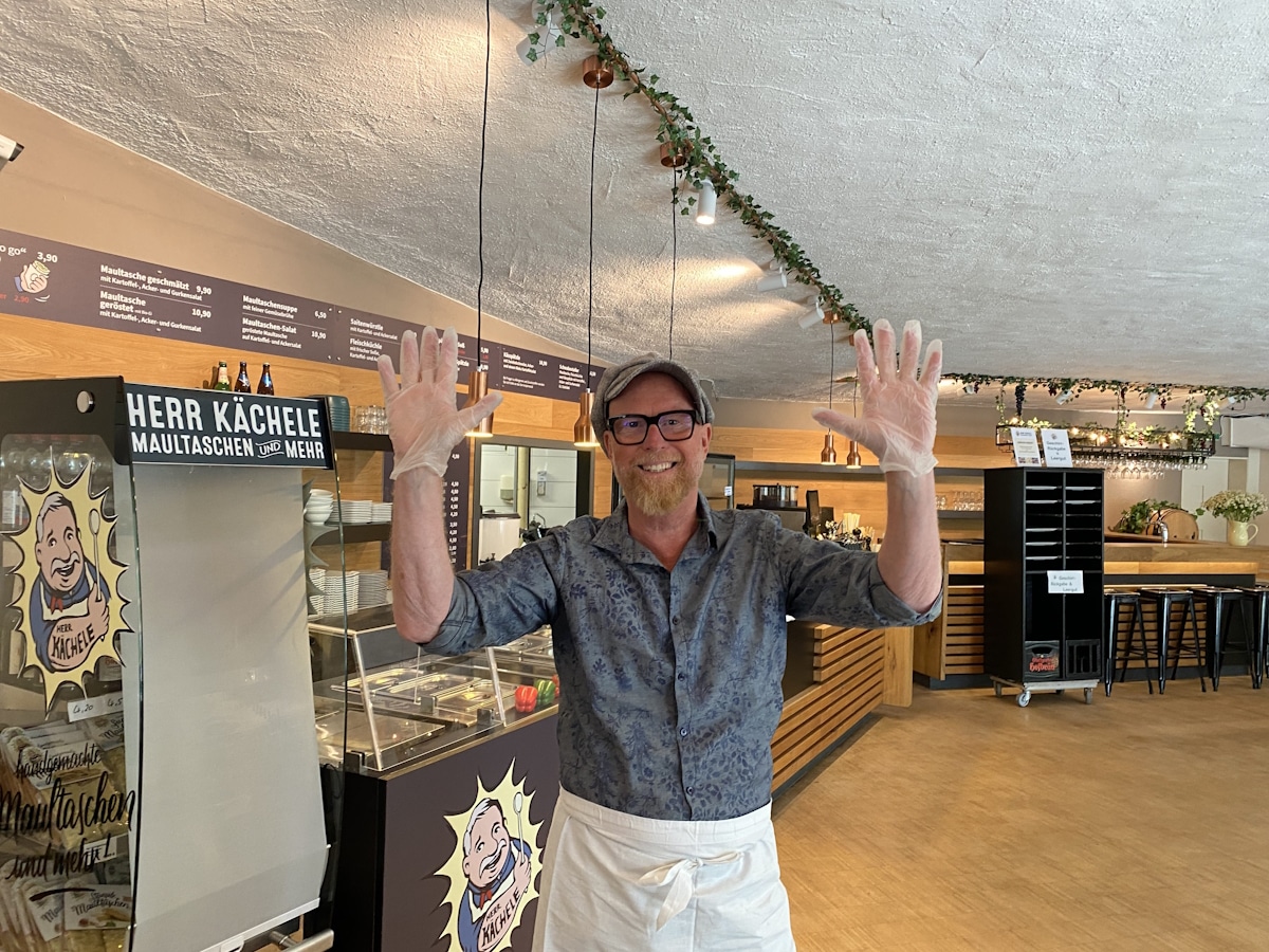 Herr Kächele 1 Innenbereich des Herr Kächele Imbiss, im Vordergrund Herr Kächele mit Schürze und Handschuhen, mit erhobenen Händen und lächelnd.