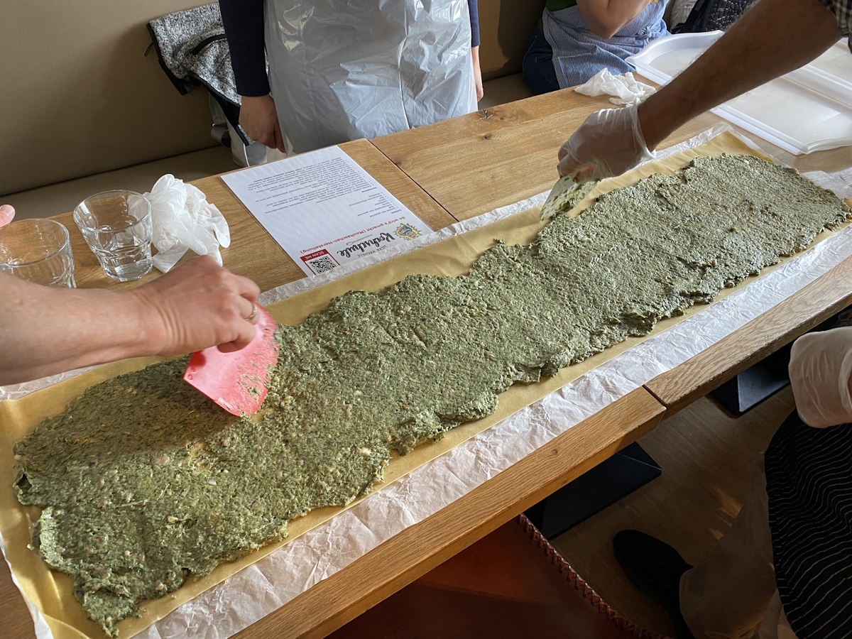 Kochkurs 1 Große Arbeitsfläche mit ausgerolltem Teig, der mit einer grünen Füllung bestrichen wird. Mehrere Hände verteilen die Füllung mithilfe von Teigschabern.