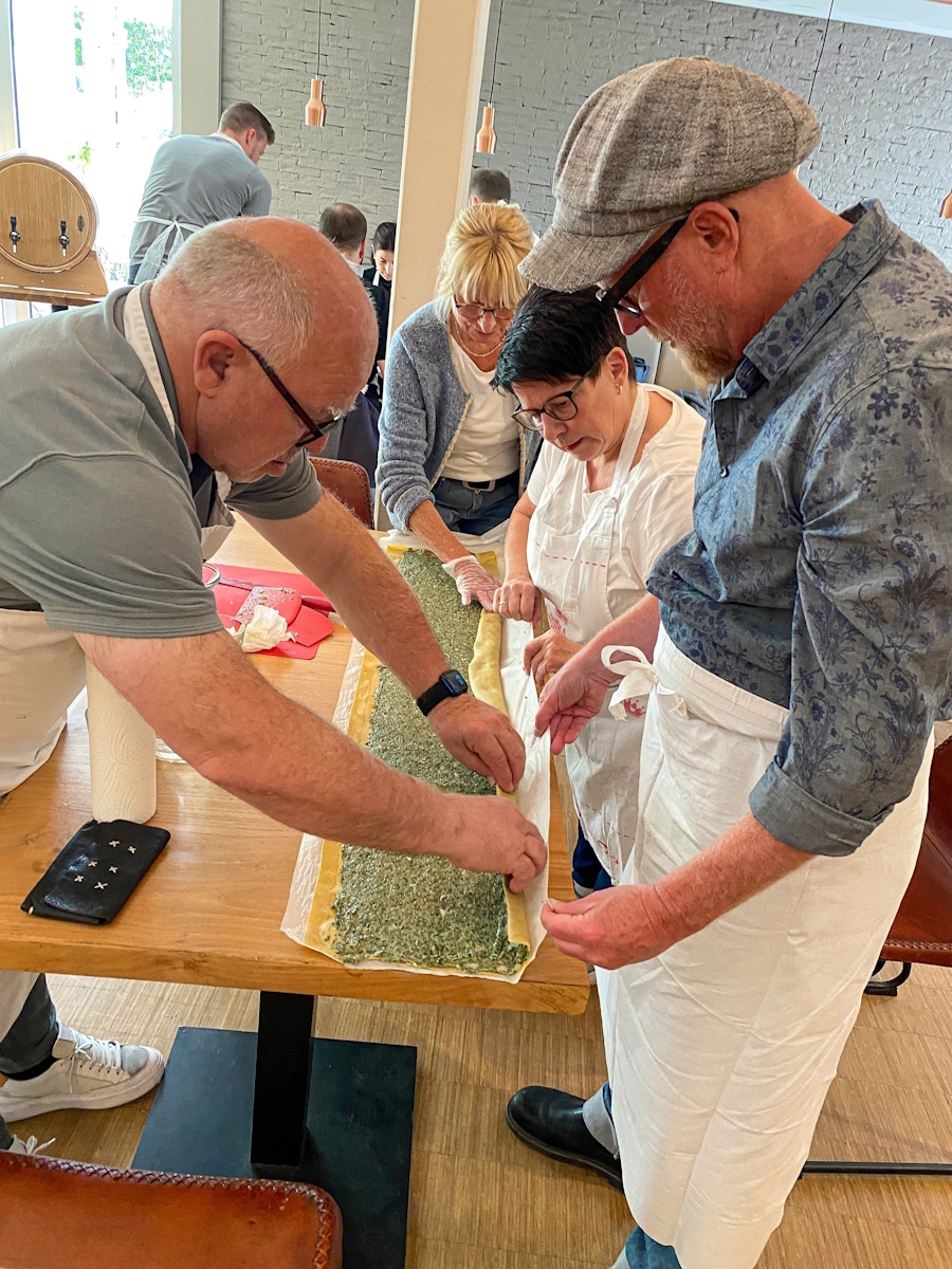 Kochkurs 3 Mehrere Personen arbeiten gemeinsam an einem langen Teigstreifen mit Füllung. Sie falten den Teig mithilfe eines Stofftuchs.