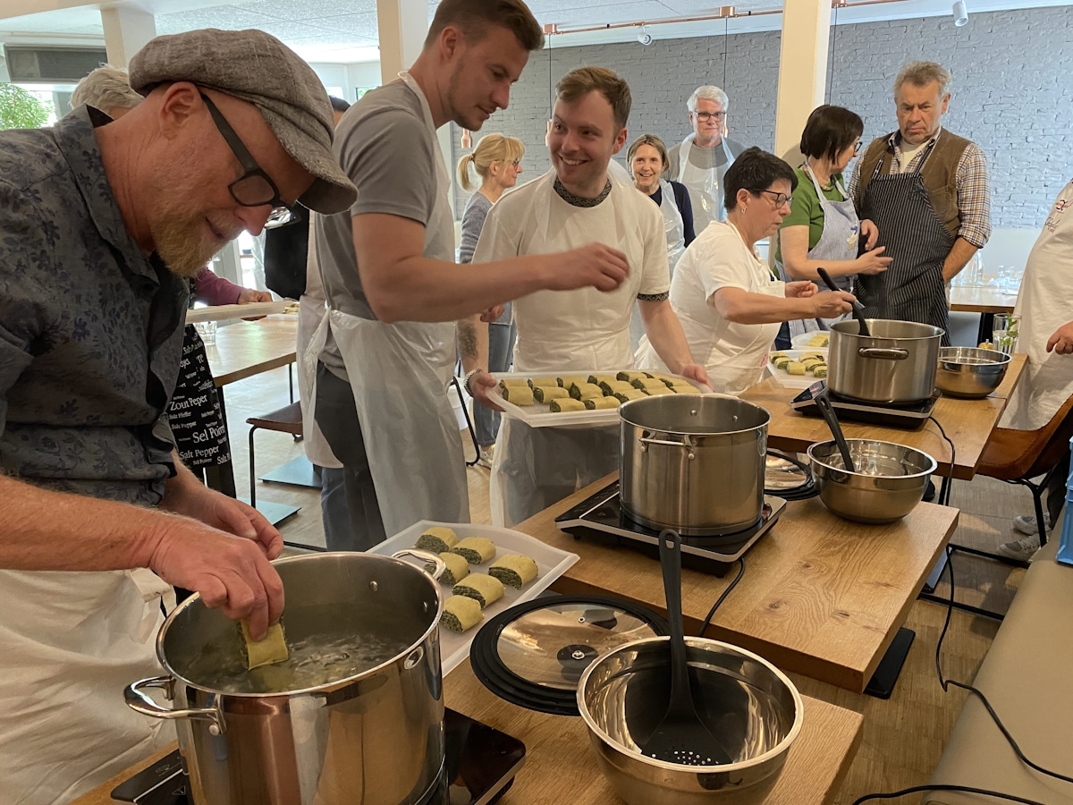 Kochkurs 4 Mehrere Personen stehen bei dem Kochkurs vor einem Tisch mit großen Kochtöpfen, Schüsseln und Tabletts mit Maultaschen. Einige Teilnehmer geben die Maultaschen ins kochende Wasser.