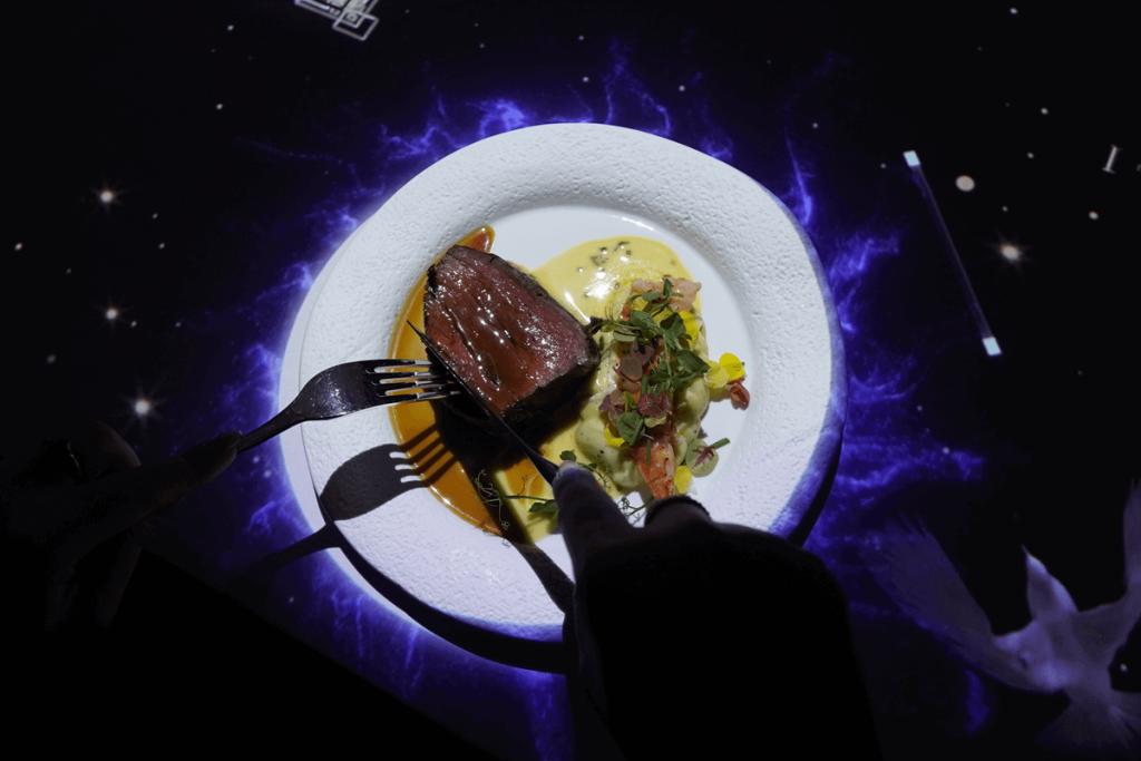 Kulinarische-Date-Ideen-The-Magic-Table - Teller mit Schwarzlich beleuchtet auf dunklem Tisch mit Steack und Soße und Gemüse und 2 Händen mit Messer und Gabel