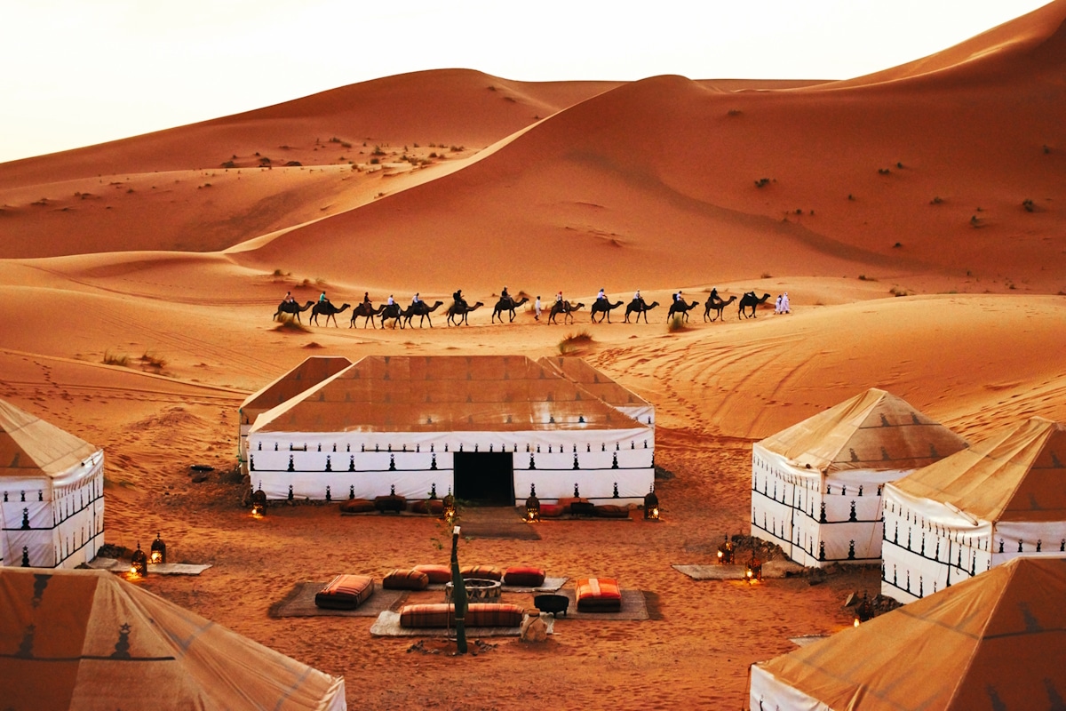 MA_Merzouga_Desert Camp Bouchedor2_HO_Desert Camp Bouchedor_Bunch of Sox, Arkaitz Latorre & Pablo de Val s Wüstenlager mit mehreren weißen Zelten, Teppichen und Sitzbereich in der Mitte, umgeben von hohen Sanddünen. Im Hintergrund reitet eine Karawane von Kamelen in einer Linie über die Dünen.