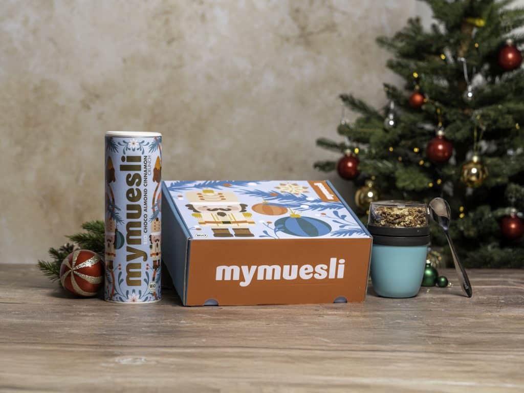 Im Vordergrund Mymuesli-Geschenkset mit Dose, Box, Becher und Löffel, dekoriert mit Christbaumkugeln und im Hintergrund ein Weihnachtsbaum.