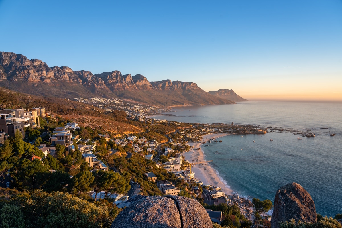 The,Rock,Viewpoint,In,Cape,Town,Over,Camps,Bay,,View Panoramablick auf die Küste von Camps Bay in Kapstadt mit weißen Sandstränden, türkisfarbenem Meer und den markanten Bergen der Zwölf Apostel im Hintergrund bei klarem Himmel.