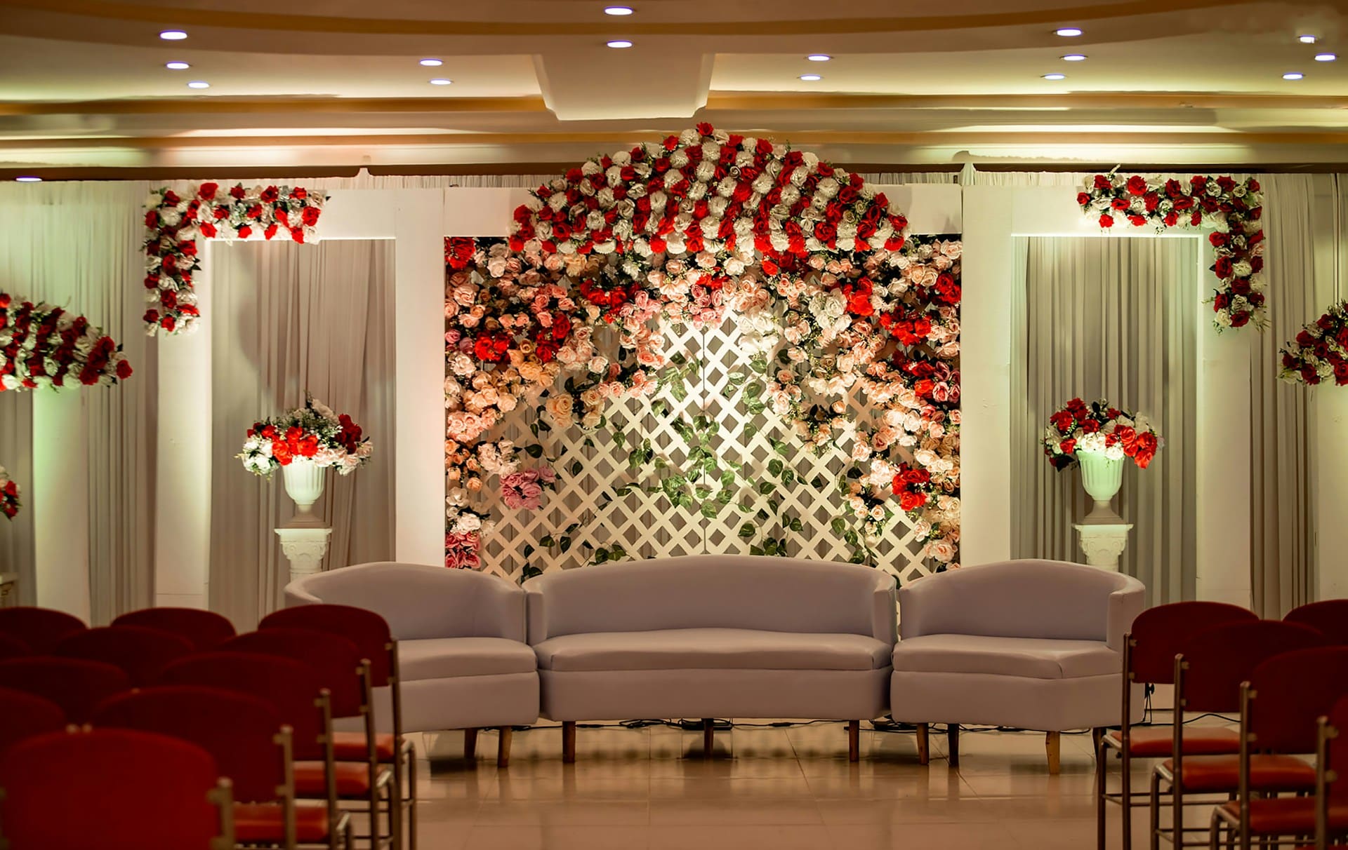 Festlich dekorierte Bühne mit weißem Sofa und zwei Sesseln. Dahinter ein weißes Gitter mit üppigem Blumenschmuck in Rot, Rosa und Weiß, arrangiert in Bögen und Vasen. Im Vordergrund sind Reihen roter Stühle sichtbar.