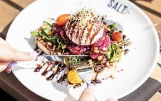 Weißer Teller mit "SALT" Logo und Toast mit Salat und Mozzarellakäse, der von 2 Händen mit Messer und Gabel zerteilt wird.