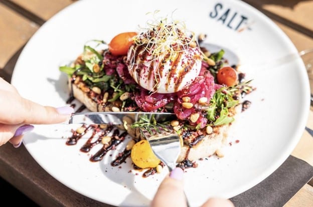 Weißer Teller mit "SALT" Logo und Toast mit Salat und Mozzarellakäse, der von 2 Händen mit Messer und Gabel zerteilt wird.