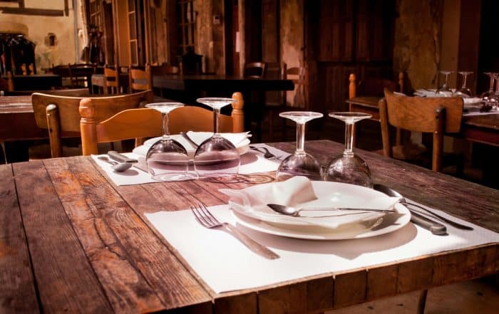 eingedeckter rustikaler Holztisch mit weißen Tischsets, weißen Tellern, Besteck und auf den Kopf gesetellten Gläsern in rustikalem und gemütlichem Ambeiten