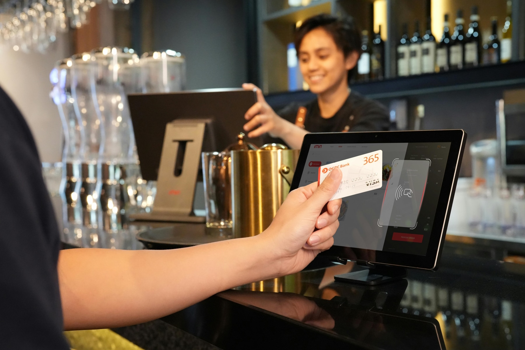 Hand mit Kreditkarte bezahlt kontaktlos am Tresen eines Restaurants. Hinter der Bar ist eine Frau am Computermonitor.