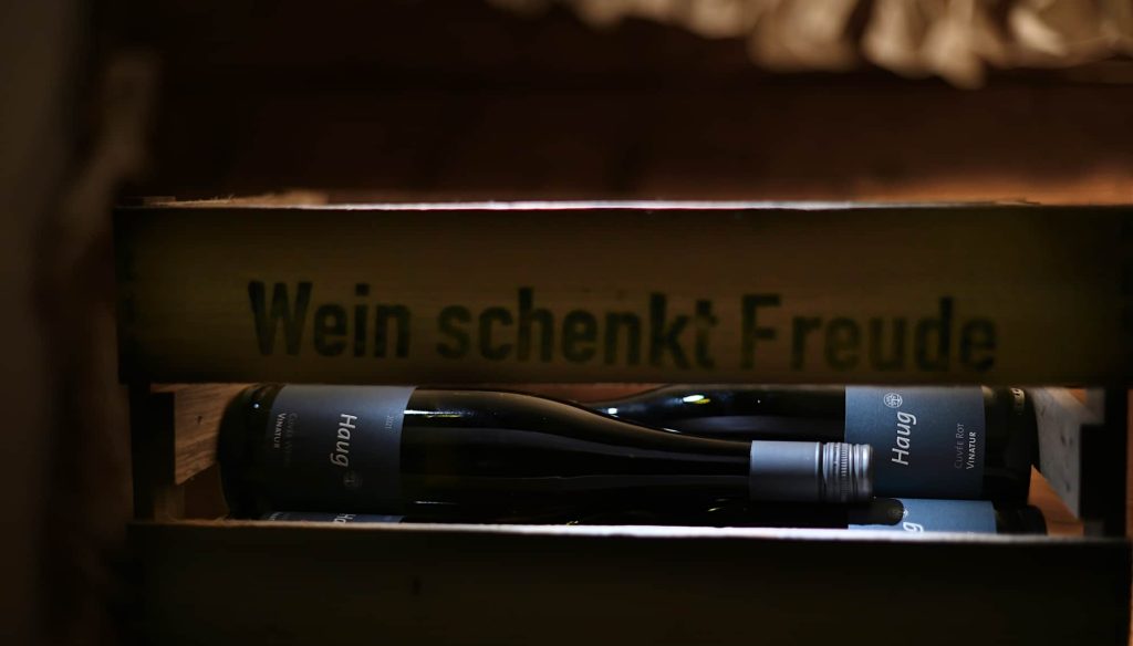 2 Weinflaschen des Weinguts Haug liegen in einer Holzkiste auf der Steht - Wein schenkt Freude