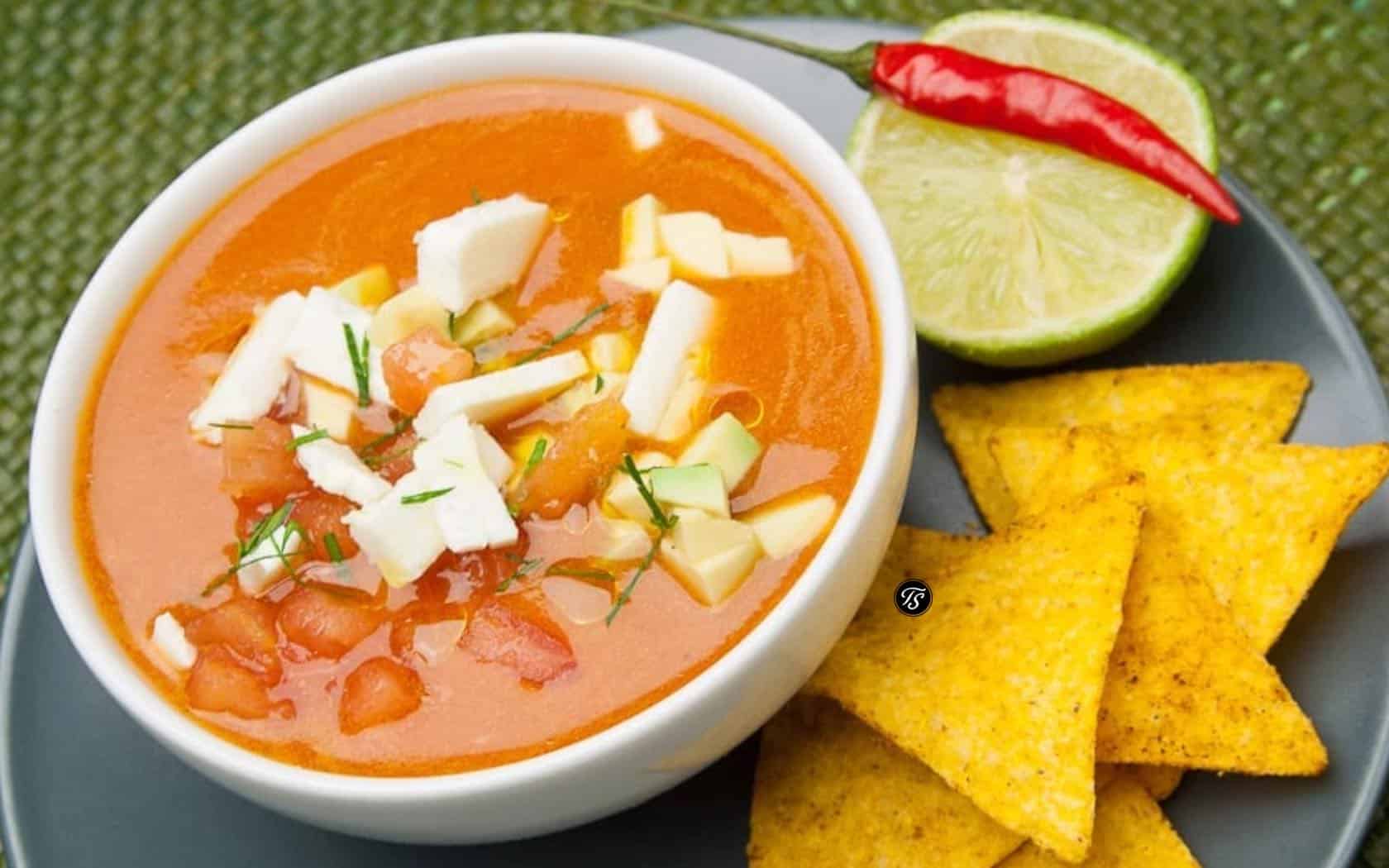 kalte-tomatensuppe-thomas-sixt-fuer-varta alte Tomatensuppe in weißer Schale mit weißen Zwiebeln neben Tortillachips und Chillischote und Limettenscheibe