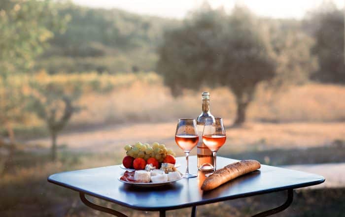 Bottle of rose wine and two full glasses of wine on table in heart of Provence, France with french bread, cheese, ham, grapes and peaches with olive trees on background