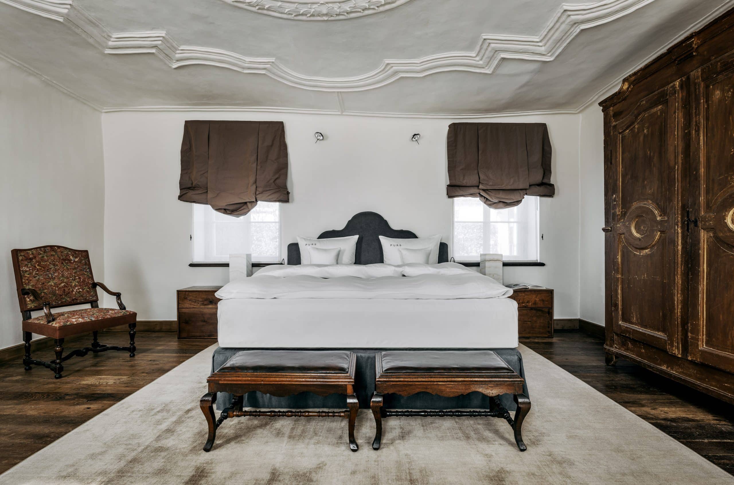 großes Bett mit weißer Bettwäsche auf dunklem Holzboden mit antikem Schrank aus dunklem Holz, Stuckdecke in der Signature Suite im PURS in Andernach