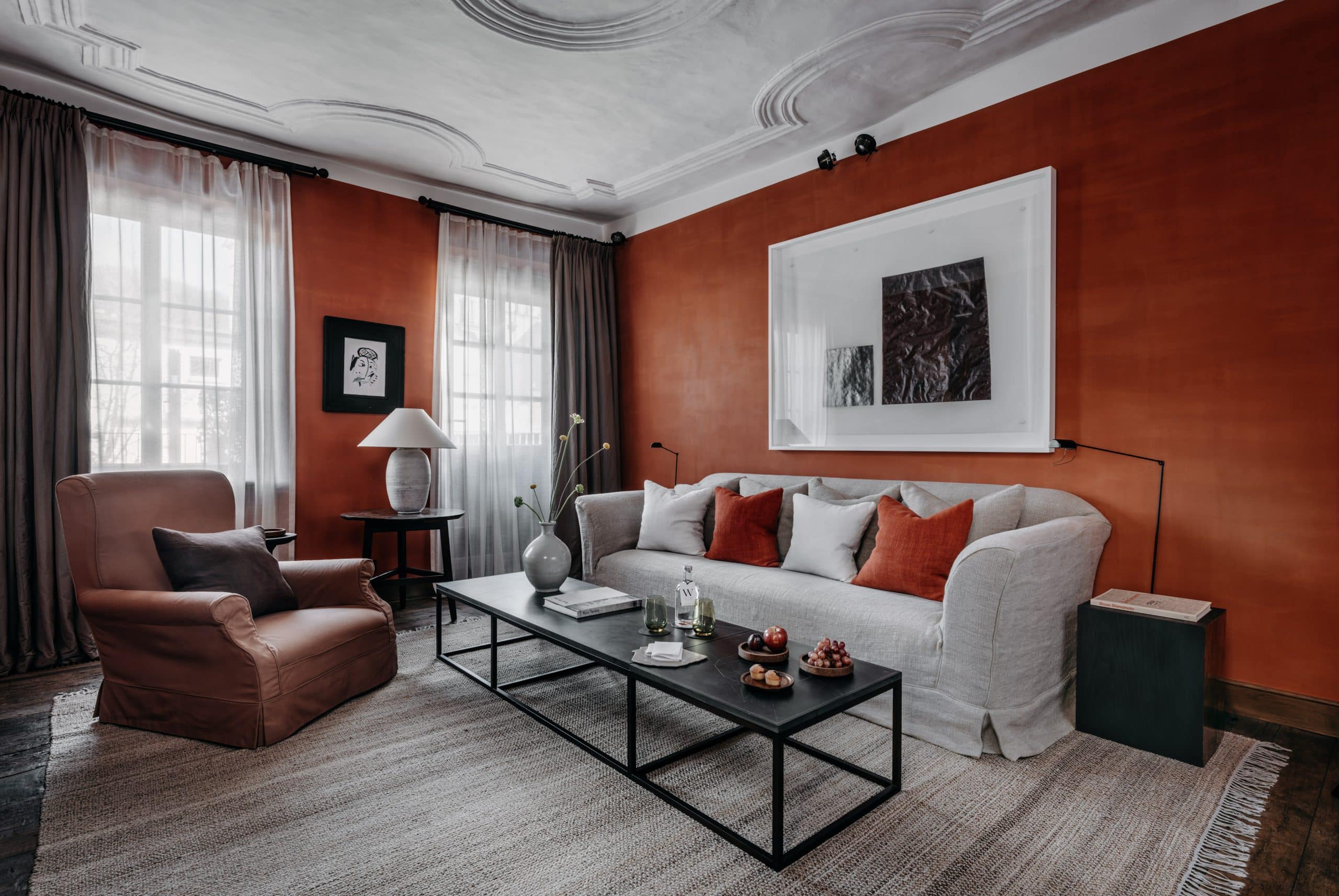 Signature Suite mit roter Wand, weißen Stuckdecken Sprossenfenster mit bodentiefen Vorhängen, Glastisch, Lampe und weißem Sofa in der Alten Kanzlei im PURS Andernach