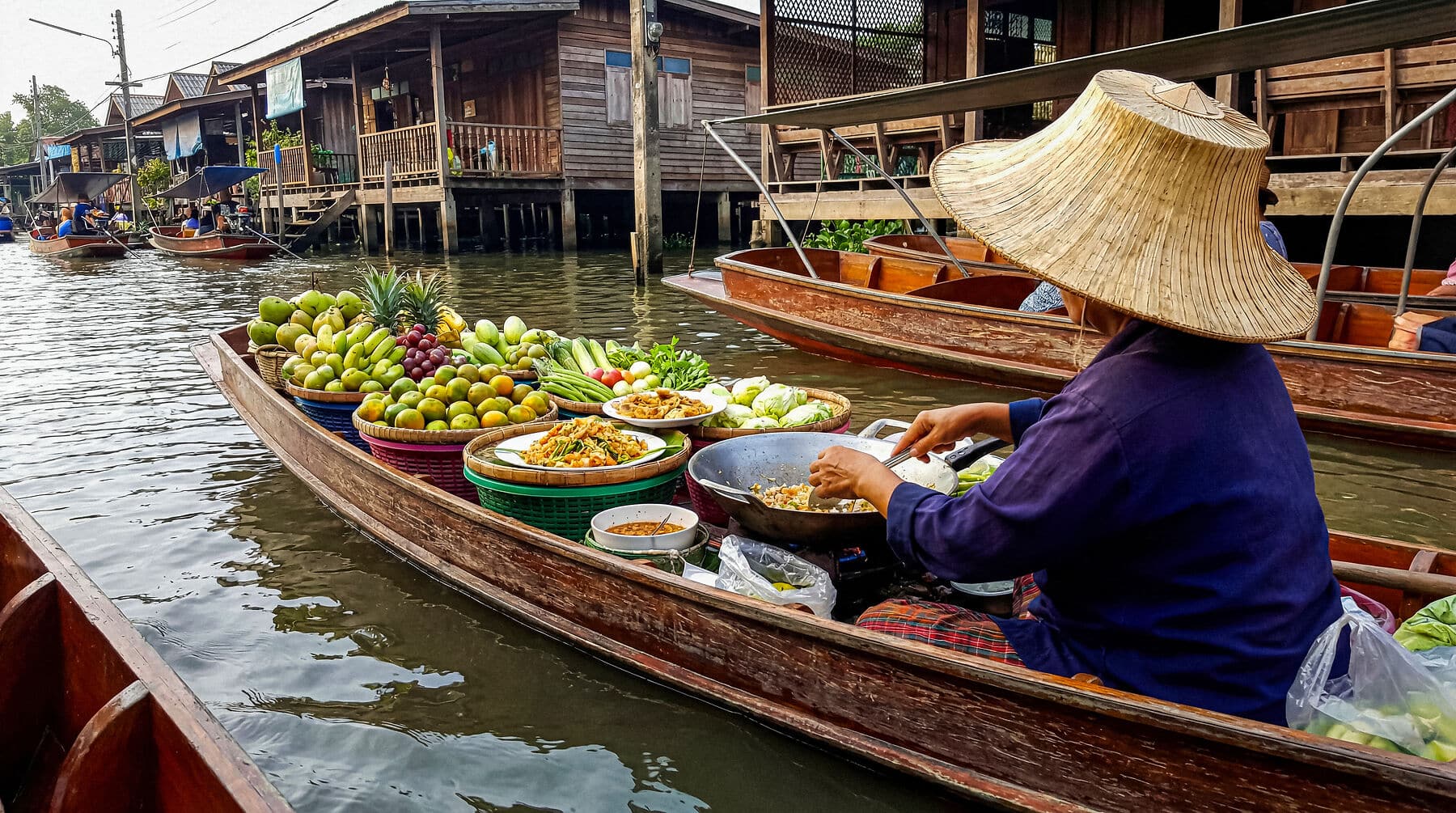 Woman in traditional hat cooks meal on boat at Thai floating market. Fresh produce. Authentic cultural scene. Ideal for travel, food.