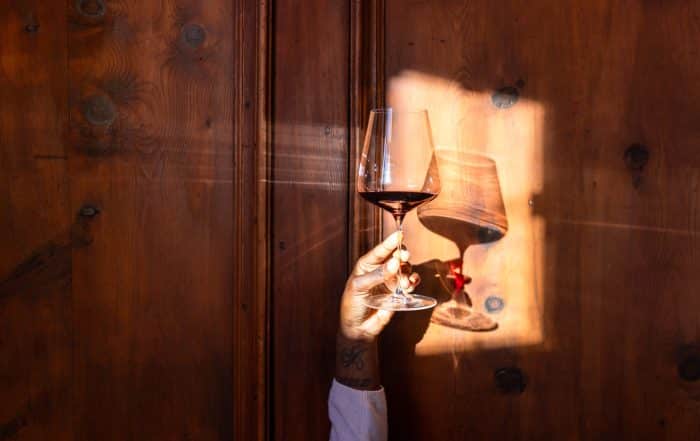 Mann hält Rotweinglas in die Luft, Detailaufnahme von der Hand und dem Glas vor holzvertäfelten Wand