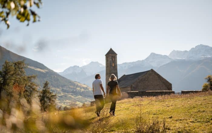 Mann und Frau laufen Hand in Hand durch die Herbstlandschaft im Vinschgau mit Kirche und Bergpanorama