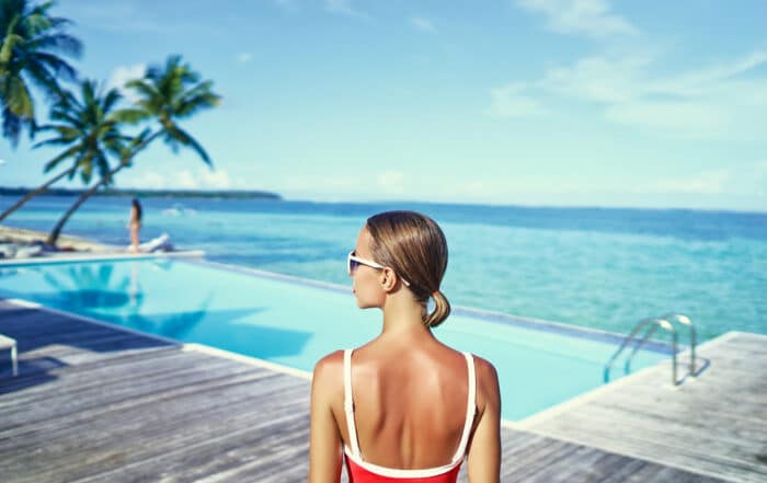 Detailaufnahme einer Frau in klassischem roten Badeanzug und weißer Sonnenbrille, die vor einem infinity-Pool am Strand steht und zur Seite blickt.