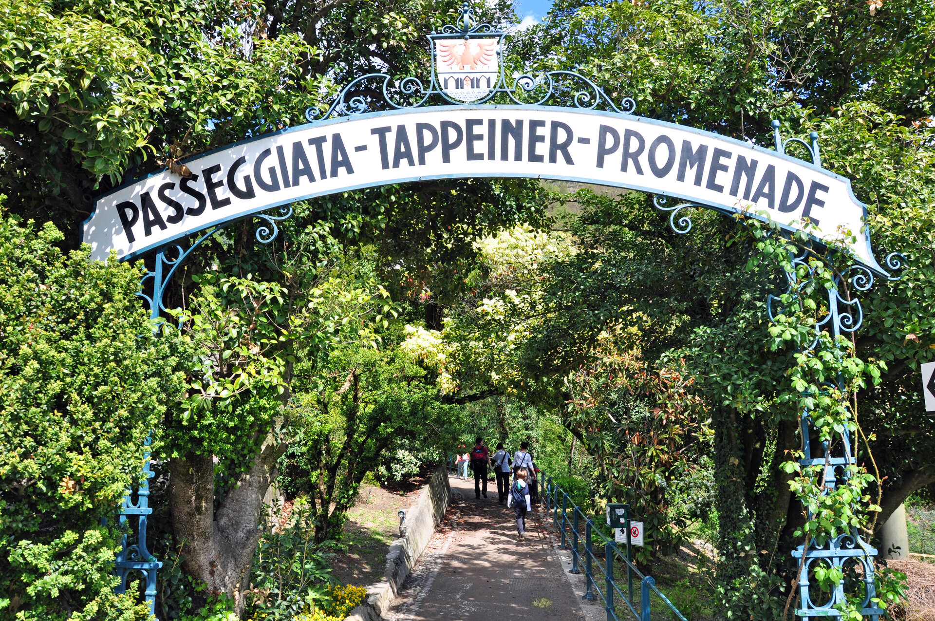 Rundes Bogenschild über Weg mit grünen Bäumen gesäumt mit der Aufschrift Passegiata-Tappeiner-Promenade